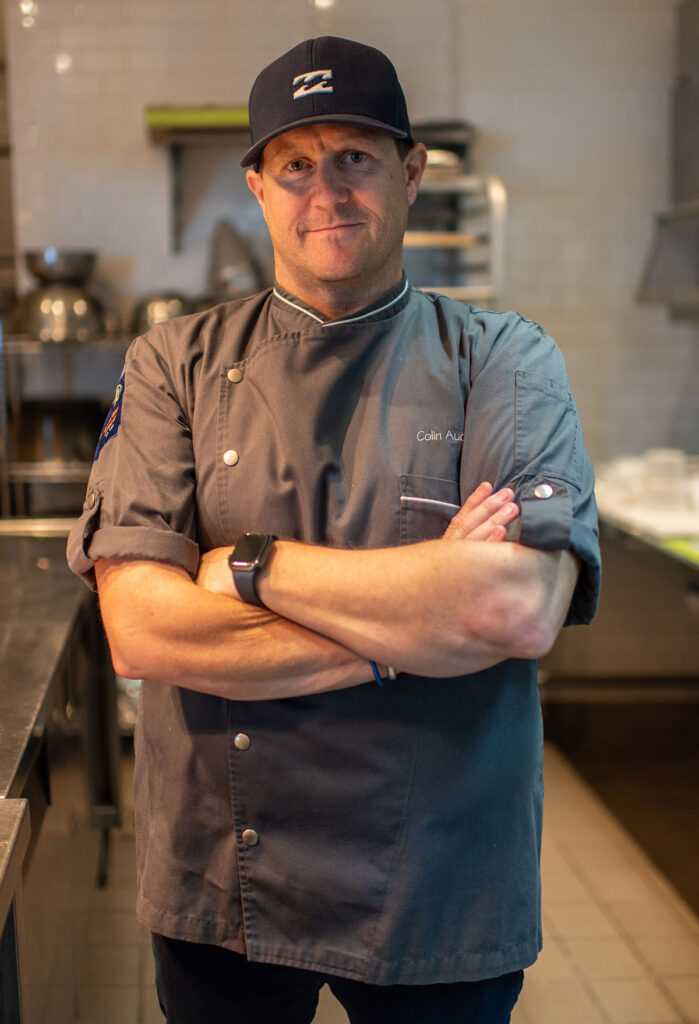 Chef Colin Auchincloss in a charcoal chef's jacket and cap, arms crossed, in a restaurant kitchen